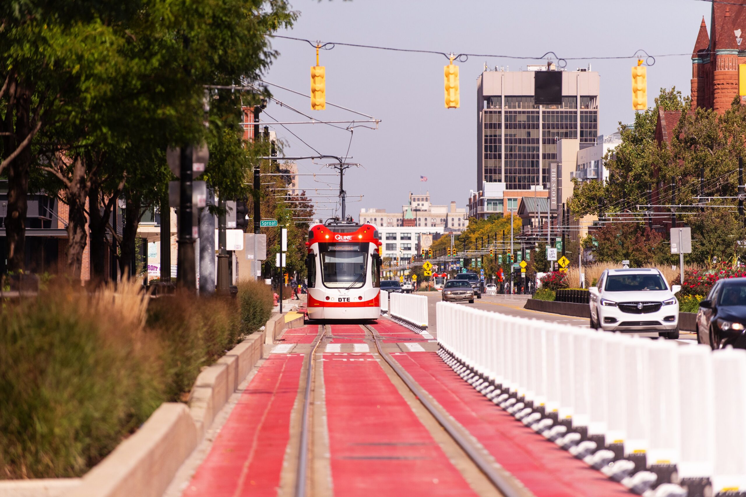 A streetcar travels along dedicated red tracks in the middle of a city street, surrounded by trees, cars, buildings, and traffic lights on a clear day.