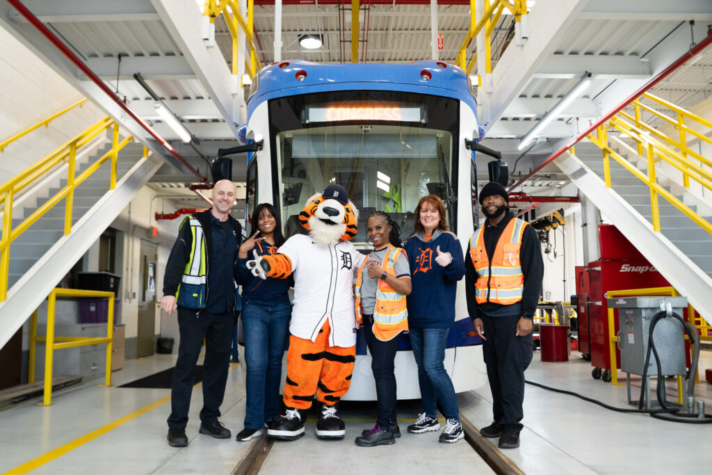 Bus service workers, pedestrians wearing Detroit gear, and a Detroit tiger mascot pose in front of a bus